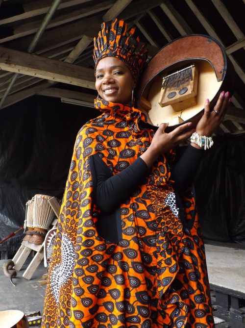 Anna Mudeka, a woman with dark skin, stands smiling at the camera and holding a Kalimba. She wears an orange patterned robe and matching hat.