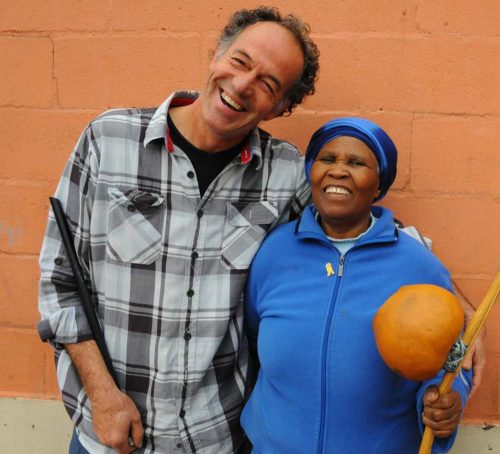Pedro Espi-Sanchis, a pale skinned man with brown and grey hair, stands next to a dark skinned woman holding. Pedro wears a grey check shirt and the woman is dressed in a bright blue jacket and head wrap. Both smile widely.