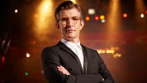 Gareth Malone stands in front of an arena that is lit to look golden. He has short brown hair, glasses and wears a tuxedo with no bowtie.